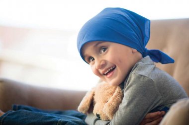 Child cancer patient wearing bandana, smiling holding a teddy bear